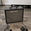 Černý kytarový kombo zesilovač s šedým textilním krytem reproduktoru a nápisem Blackstar. K zesilovači je připojen černý nožní spínač s logem Blackst…