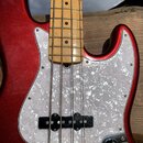 Detail elektrické basy s červeným metalizovaným povrchem korpusu. Nástroj má bílý pickguard s perleťovým vzorem a hmatník z jasného dřeva s černými p…