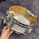 Fotografie znázorňuje malý bílý buben s chromovaným obručováním a mechanismem na straně. Na horní bláně je vidět logo AQUARIAN, nápis PERFORMANCE SER…