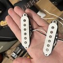 Dvě elektrické snímače typu single-coil s bílými plastovými kryty a černým základem. Na obou krytech je od ruky napsaný nečitelný text. Snímače mají…