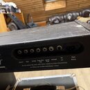 Zadní strana černého elektronického zařízení v kovovém šasi s viditelným opotřebením povrchu a škrábancemi. Na zadním panelu se nachází řada konektor…