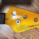 Detail hlavy basové kytary z žlutého dřeva se čtyřmi otvory na ladičky. Na povrchu je černý nápis Vintage ICON Series™ a v pravém dolním rohu malý ov…