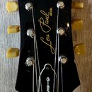 Černá hlava elektrické kytary s bílým logem Epiphone v horní části. Uprostřed je zlatý nápis Les Paul MODEL. Na krytu ladicího mechanismu je svisle n…