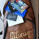 Hnědý kytarový pouzdro z imitace kůže s velkým zlatým nápisem Gibson. Na pouzdře leží modrá karta s nápisem GIBSON WORLD WARRANTY, černý utěrník s bí…