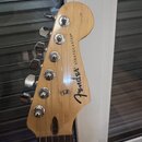 Fotografie zachycuje hlavu kytary z přírodního dřeva s chromovanými ladičkami. Na dřevě je černý nápis Fender a STRATOCASTER®. Povrch dřeva vykazuje…