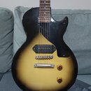 Elektrická kytara s tělem v žlutém gradientu (sunburst) a černým pickguardem. Nástroj má jeden humbucker v pozici mostu, dva zlaté ovládací knofíky a…