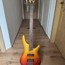 Elektrická basová kytara s tělem v žlutě-červeném přechodovém odstínu (sunburst) a hnedým hmatníkem. Na hlavě nástroje je červený nápis "Saga". Kytar…