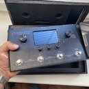 Šedý efektový procesor s kovovým povrchem a displejem uprostřed. Na zařízení je nápis AMPERO Amp Modeler / Effects Processor a čtyři stomečkové spína…