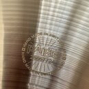 Detail kovového povrchu činely s koncentrickými rýhami. Uprostřed je vyražený nápis PAISTE, pod kterým je sériové číslo 25057724. Celý nápis je obklo…