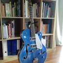 Modrá polodublová elektrická kytara s f-otvory a bílým pickguardem. Na hlavě hрифu je nápis GRETSCH a na pickguardu je viditelné logo GRETSCH. Nástro…