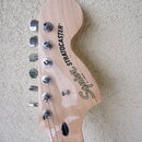 Fotografie zachycuje hlavu kytary z přírodního světlého dřeva s chromovanými ladičkami. Na povrchu je vidět nápis Squier STRATOCASTER® a menší text B…