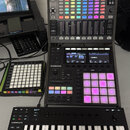 Na fotografii jsou tři šedé kontrolery značky Native Instruments. V horní části je zařízení s nápisem MASCHINE JAM, které obsahuje mřížku podsvícenýc…