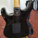 Zadní strana elektrické kytary s černým glossy povrchem a světle hnědým dřevěným hmatníkem. Na kovové spojce krku je nápis „Fender, Inc. MADE IN USA“…