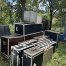 Skupina transportních case různých velikostí a barev (černé, hnědé, stříbrné) umístěná na trávníku. Na jedné z černých case je viditelný nápis ACCUSR…