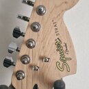 Fotografie zachycuje hlavu elektrické kytary z přírodního světlého dřeva s tmavým hmatníkem. Na hlavě je žlutý nápis Squier by Fender a černý text ST…