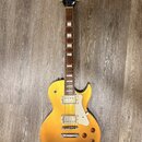 Zlatá elektrická kytara typu Les Paul s dvěma humbuckery a bílým plechtrátem. Na hlavě je vidět logo Cort. Kytara má čtyři ovládací knoflíky a hриф s…