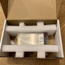 V kartonové krabici s polystyrenovou výplní je umístěn kovový předmět stříbrné barvy. Na boku je viditelný štítek s logem ETL a texty: CONFORMED TO,…