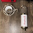 Na fotografii je stříbrný kondenzátorový mikrofon s bílým tělem a doprovodný šokabsorber v kovovém provedení. Na bílé části mikrofonu je červený nápi…