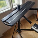 Černý kovový stojan s dlouhou, úzkou pracovní deskou. Na desce je umístěn černý cylindrický držák. V pozadí je vidět část klávesového nástroje. Povrc…