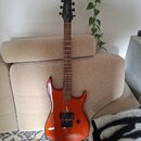 Elektrická kytara s oranžovým lakovaným tělem a hnědým hnedým hmatníkem. Na hlavě je vidět bílý nápis "Ibanez". Kytara má dva humbuckery, jeden ovlád…