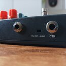 Záběr na zadní stranu černého efektpedálu s matným povrchem. Jsou zde vidět dva kovové konektory a mezi nimi nápis TAP/EXP — AUDIO a CTR. V horní čás…