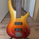 Elektrická basová kytara s tělem v červeno-žlutém přechodovém lakování (sunburst) a hnědým hmatníkem. Nástroj má dva snímače typu humbucker a čtyři o…