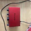 Červený kovový audio rozhraní s nápisem Focusrite na horní straně. Na přední hraně jsou vidět čtyři ovládací prvky v podobě knoflíků. Zařízení je při…