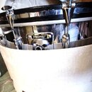 Chromovaný buben umístěný v bílém plátěném přepravním pouzdře s hnědým koženým úchem. Na kovovém šperku je viditelný nápis „Sluggerland“. Povrch bubn…