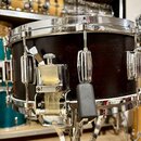 Detailní pohled na snare drum s tmavě hnědým dřevěným korpusem a chromovanými obručemi. Na levé straně je vidět mechanismus s přilepeným kusem žluté…