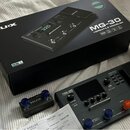 Na fotografii je kytarový procesor NUX MG-30 v černé barvě, doprovázený originální krabicí a dokumentací. Na přístroji je vidět nápis MG-30 VERSATILE…