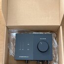 Černý audio rozhraní v kartonové krabici s plastovou fólií. Na zařízení jsou nápisy „USB“, „IN 1“, „IN 2“, „48 V“, „OUT 1/2 VOLUME“ a v dolní části l…