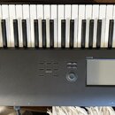 Černý digitální klavír s bílými a černými klávesami. Na levé straně korpusu je bílý nápis NAUTILUS MUSIC WORKSTATION. V pravé části se nachází disple…