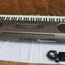 Šedý elektronický klavír s plastovým povrchem a černými reproduktory. Na přední straně je umístěn nápis CASIO. Před nástrojem leží černý síťový adapt…