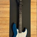 Modrá elektrická basová kytara s bílým perleťovým pickguardem. Na hlavě hmatníku je viditelné logo Fender. Nástroj má čtyři struny a kový most. Povrc…