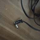 Černý kabel zakončený kovovým konektorem. Spojení kabelu a konektoru je obmotáno černou izolační páskou, která vykazuje známky opotřebení a rozlepová…