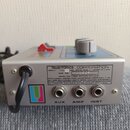 Stříbrný efektový pedál s ovládacími prvky na horní straně. Na zadním štítku je nápis musitronics CORPORATION, INPUT: 110-240 VAC, 60HZ, 5VA.. MADE I…