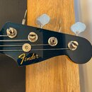 Detail hlavy elektrické basy v tmavě modré barvě. Na hlavě je žlutý nápis Fender a vedle něj text JAZZ BASS ELECTRIC BASS. Nástroj má čtyři kovové la…