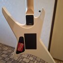 Bílá elektrická kytara s ostrými hranami korpusu. Na instrumentu jsou tři černé kryty elektroniky, z nichž jeden nese červenobílý nápis EMG. Na horní…