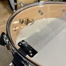 Detailní pohled na bílý buben s průhledným obručem. Na blaně je viditelný nápis EVANS CALFGRAM 1980 RESONANT SERIES 300. Přes blanu je natažen kovový…