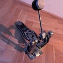 Stříbrný basový pedál s černým šlapadlem. Na šlapadle je bílý nápis TAMA a černý nápis The Liquid Co. Construction. Povrch kovových částí vykazuje zn…