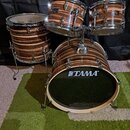 Bubnová sada s povrchem z tmavého pruhovaného dřeva. Na předním bubnu je bílý nápis TAMA®. Sada obsahuje basový buben, dva tom-tomy a jeden floor tom…