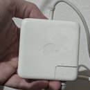 Bílý čtvercový napájecí adaptér z plastu s vyembasovaným logem Apple uprostřed. Povrch je mírně znečištěný a vykazuje známky opotřebení. Z horní část…