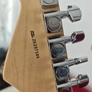 Záběr na zadní stranu dřevěné hlavy kytary s pěti chromovanými ladičkami. Na každé ladičce je vyryto logo Fender. Na dřevě je černý nápis SERIAL NUMB…