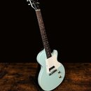 Světle modrá elektrická kytara s bílým pickguardem a dvěma humbuckery, umístěná na černém stojanu. Na hlavě hmatníku je zlatý nápis Gibson. Kytara má…