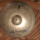 Kovový činel v přirozené zlatavé barvě s viditelným rýhováním. V horní části je černý nápis „alchemy cymbals“. Ve spodní části je černý kříž a vedle…