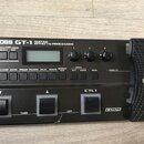 Černý kytarový procesor s nápisem BOSS GT-1 GUITAR EFFECTS PROCESSOR. Zařízení obsahuje displej, tři ovládací knoflíky, řadu tlačítek a dvastředý ped…