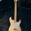 Elektrická kytara s bílým tělem a světlou hnedou hmatníkem. Na hlavě je viditelné logo Fender. Kytara má perleťový pickguard a zlaté kování ladících…