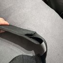 Černý popruh z textiledního materiálu s výraznou strukturou. V horní části je viditelné roztržení látky. Popruh leží na šedém povrchu.