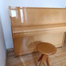 Světle hnědé dřevěné piano s glossy povrchem a dopřሁንěným dřevěným taburetem. Nástroj je umístěn u bílé stěny na dřevěné podlaze. Na viditelných část…