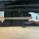 Elektrická basová kytara s tělem v sunburst provedení a bílým pickguardem, umístěná v černém tvrdém kufru. Na hlavě nástroje je viditelné logo Fender…